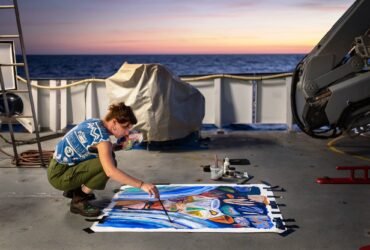 artist Ellie Hannon working on a painting on the aft deck of a research vessel at sunset