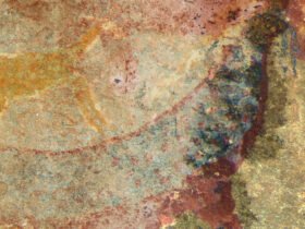 Faded paint on a cave wall appears to show two creatures, one in yellow top left and one in more reds and blues, that create an animal with an elongated body and what appears to be tusks, in blue, near its head region.