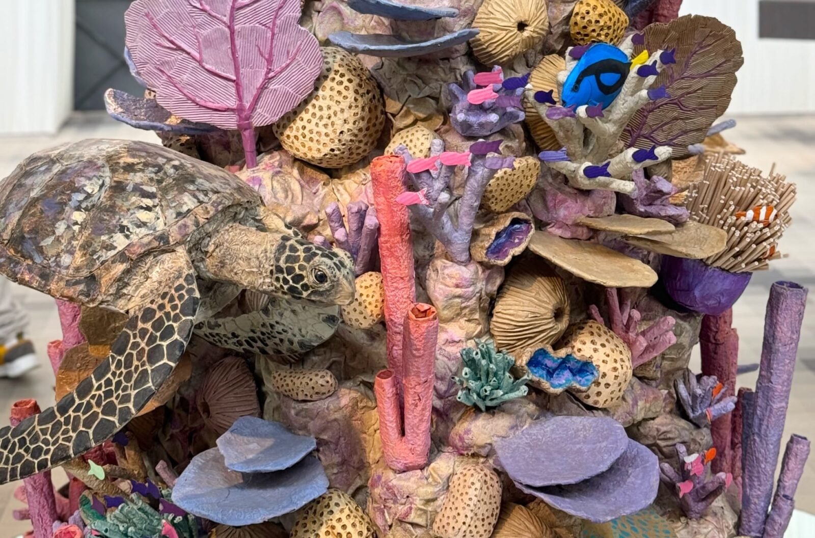 a detail of a large cardboard sculpture of a coral reef, focusing on a sea turtle and numerous small fish