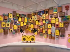 a collection of speakers painted with yellow and brown faces against a pink wall. a gramophone and boombox painted similarly stand in the front