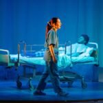 On stage, a woman in scrubs walks past a man in a hospital bed