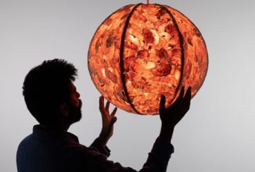 a male figure, silhouetted, reaches out to hold a glowing pendant lamp with a shade made out of sewn-together orange peels