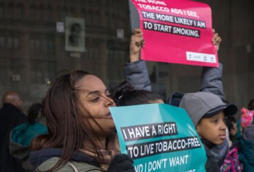 Two young people hold anti-tobacco signs at a a rally. The girl in front holds an teal-colored sign that reads, "I have a right to live tobacco-free." Another person holds a red sign that says, "The more tobacco ads I see, the more likely I am to start smoking."