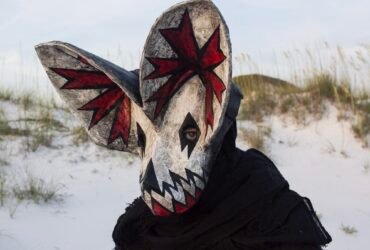 a portrait of a person wearing an ornate papier mache bat mask with red paint in the ears in a coastal setting