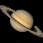 This image shows Saturn, ringed and in tones of tan, against an inky background. Three tiny light spots in the background and one dot against the lower half of the planet are four of Saturn