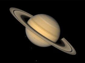 This image shows Saturn, ringed and in tones of tan, against an inky background. Three tiny light spots in the background and one dot against the lower half of the planet are four of Saturn