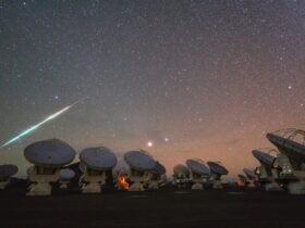 A bright light streaks across a starry, rosy-purple night sky on the left side of this image. An array of many radio telescopes are seen in the foreground.