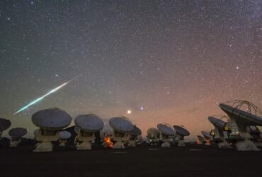 A bright light streaks across a starry, rosy-purple night sky on the left side of this image. An array of many radio telescopes are seen in the foreground.