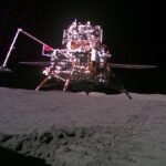 A shiny robotic lunar lander sits on a gray surface with a black background.