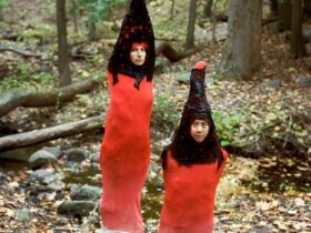 in the forest, two figures wear matching red garments reminiscent of dog stinkhorn mushrooms.