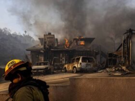 Firefighter in the foreground walks by a burning house in the background.