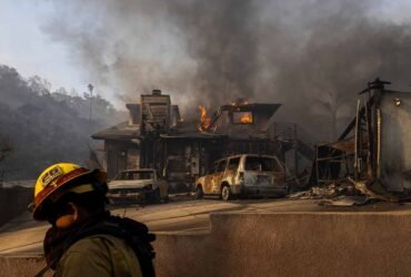 Firefighter in the foreground walks by a burning house in the background.