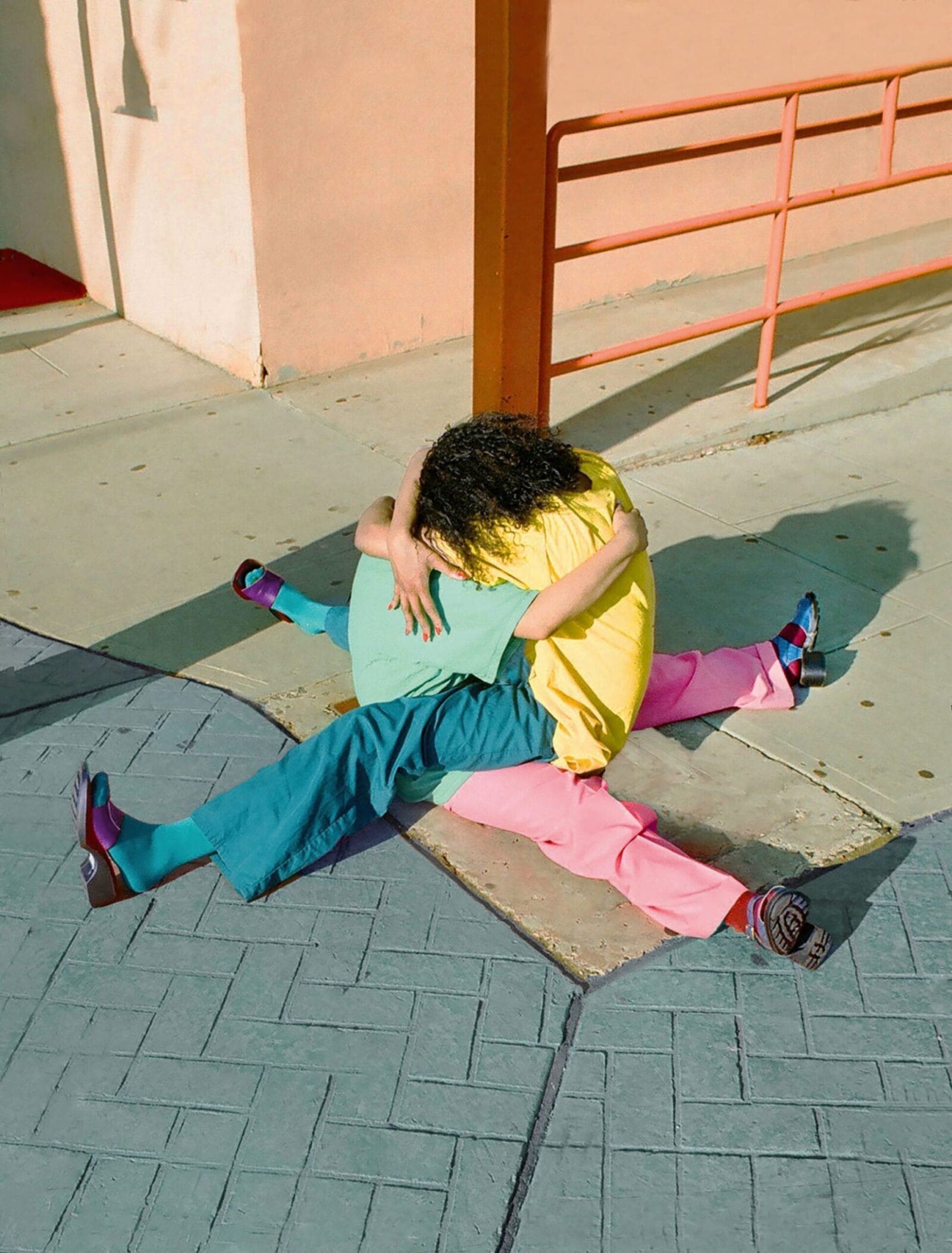 two people, one in a blue shirt and pink pants and the other in a yellow shirt and blue pants, embrace while seated on the sidewalk