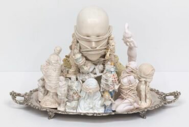 a sculpture made from found objects and figurines tied together with string atop a silver serving platter