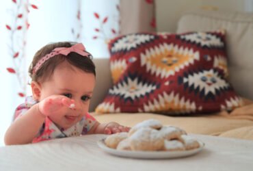 A little girl stands by a table reaching for a plate full of cookies.