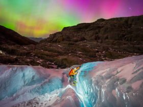 a glacier hiker climbs in the foreground with a headlamp as a rainbow colored aurora dances above