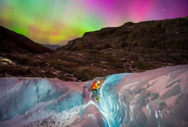 a glacier hiker climbs in the foreground with a headlamp as a rainbow colored aurora dances above