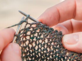 A close-up view of fingertips holding silver knitting needles, with light shining through the loose stitches in the cast fabric