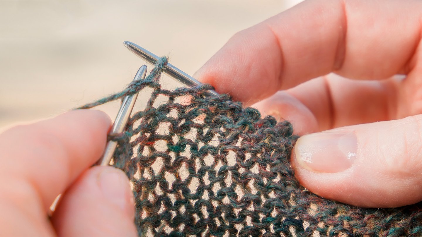 A close-up view of fingertips holding silver knitting needles, with light shining through the loose stitches in the cast fabric