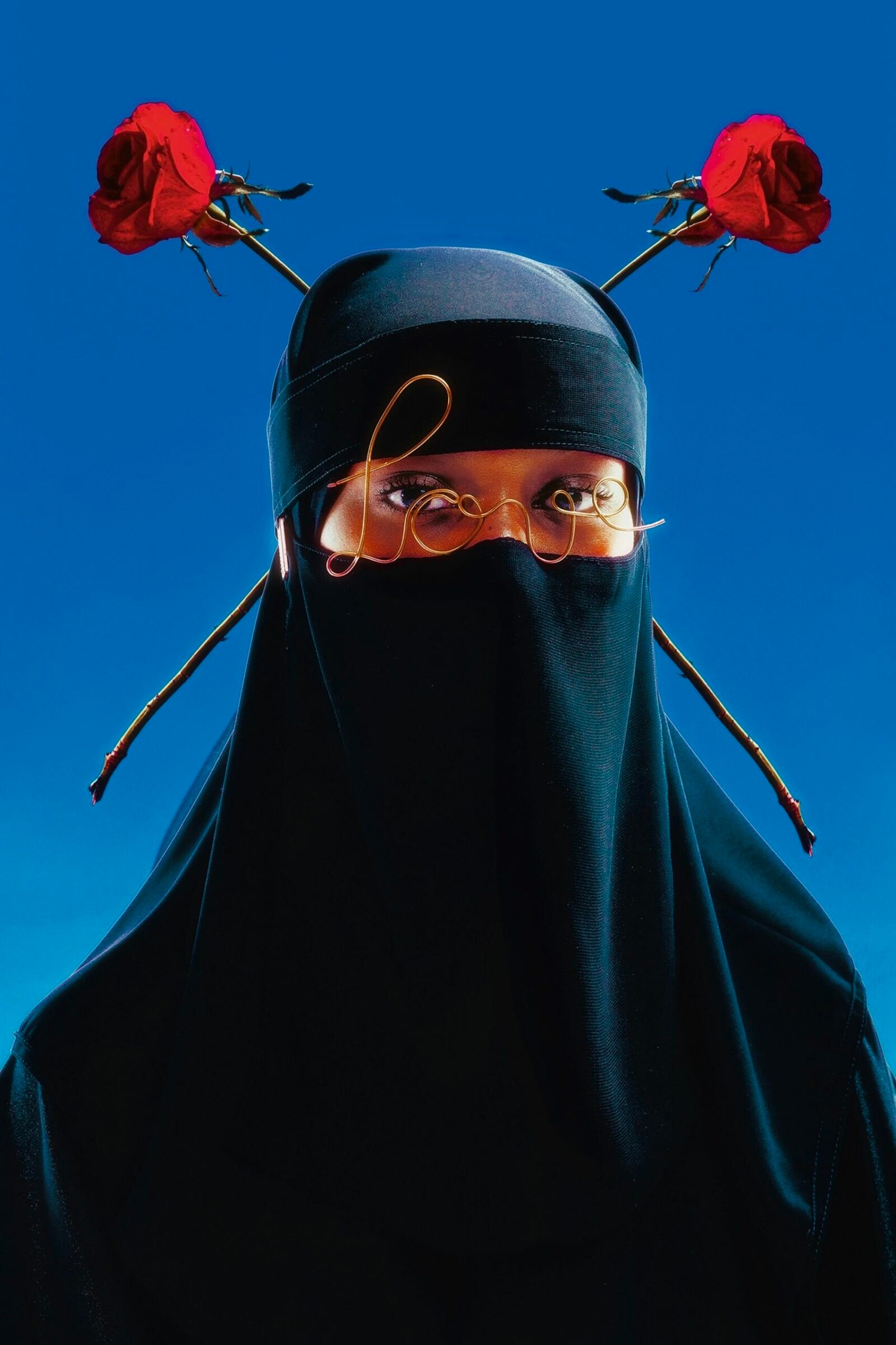 a striking portrait of a figure wearing a niqab, with roses on her head and wire worn like glasses in front of her eyes that reads "love"