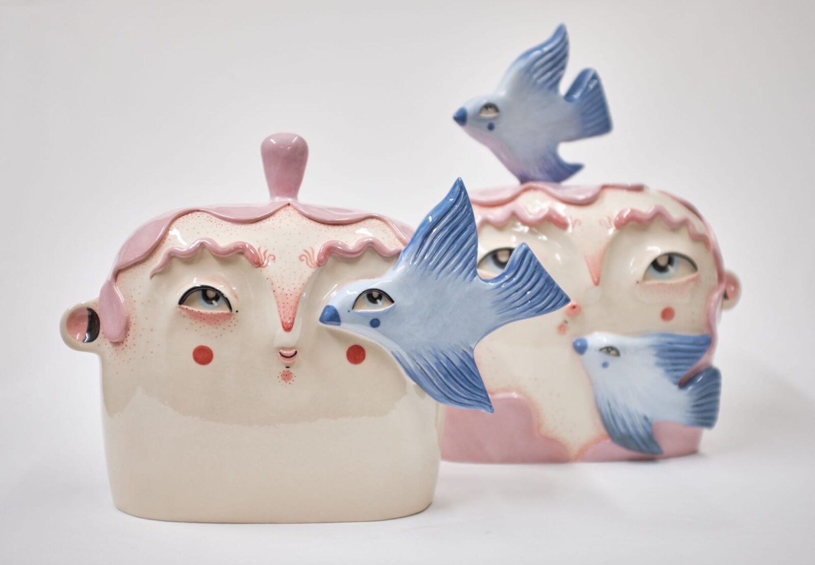 two ceramic sculptures of cute heads and faces with pink hair, with bluebirds flying around them