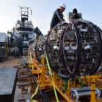 Workers on a ship prepare to launch a giant round piece of equipment off of a bright yellow ramp