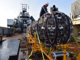 Workers on a ship prepare to launch a giant round piece of equipment off of a bright yellow ramp