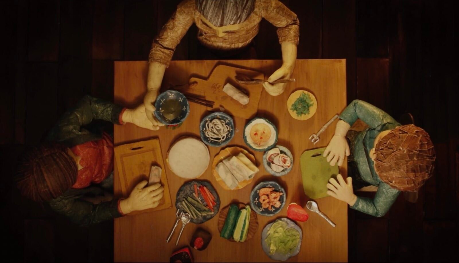 a screenshot from a stop-animated film short of an aerial view of a family around a table making spring rolls