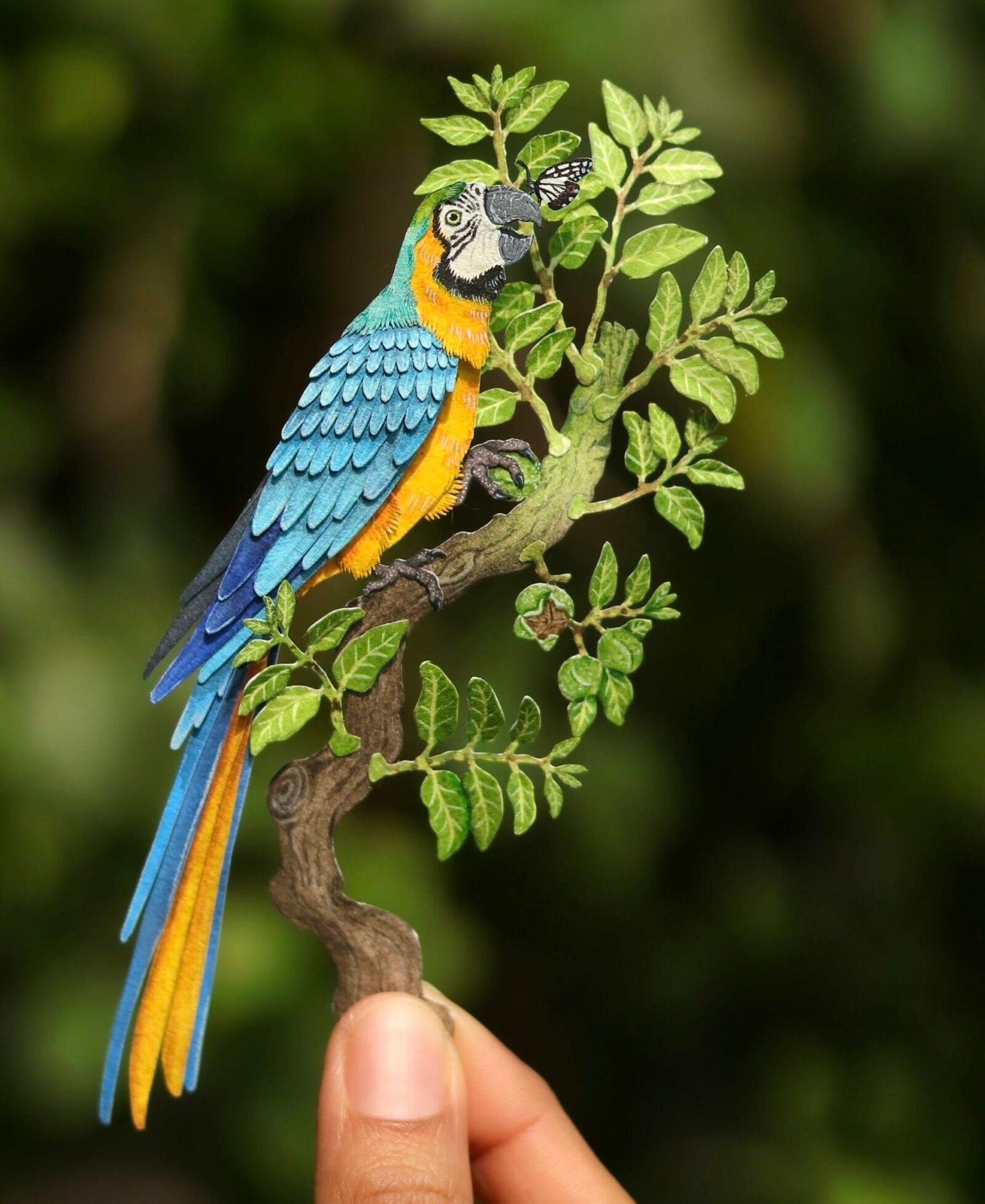 paper cutout artworks of a macaw sitting on a branch, held by the artist's hand to show tiny detail