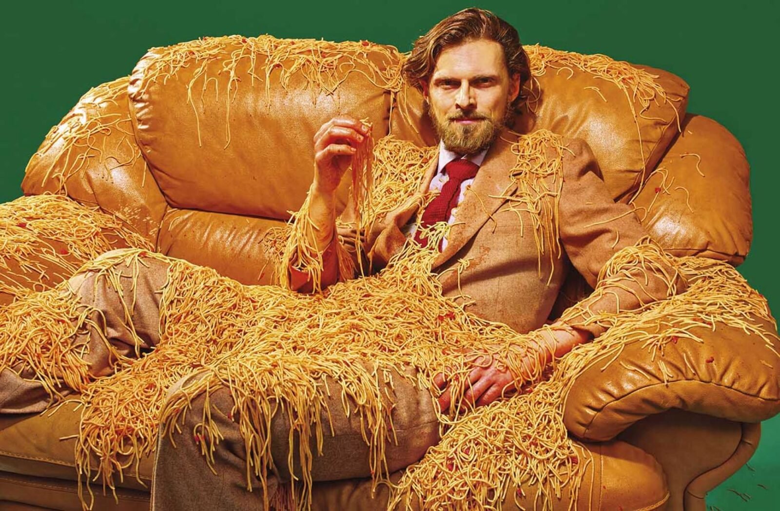 a portrait of a man in a brown suit, sitting on a brown leather couch, covered in spaghetti