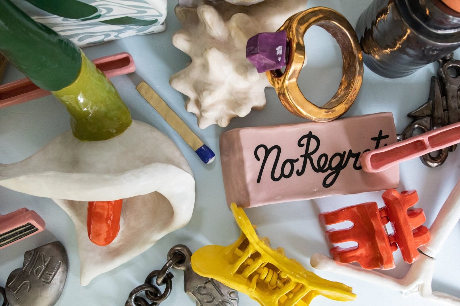 a scattered assemblage of ceramic sculptures including a flower, ring, seashell, eraser that says "no regrets," claw clips, jewelry chains, lipstick, a matchstick, disposable razors, and an IUD.