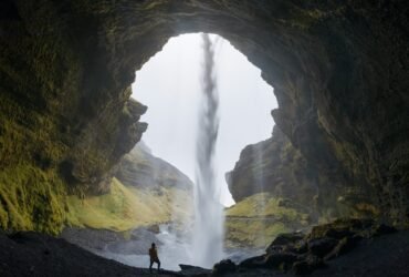 a figure stands in the opening of a cave, its silhouette accentuated against a waterfall