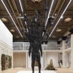 a large-scale bronze figure with branches and leaves for a head stands in a gallery