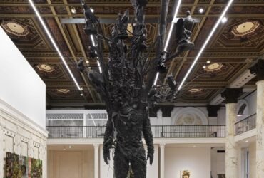 a large-scale bronze figure with branches and leaves for a head stands in a gallery