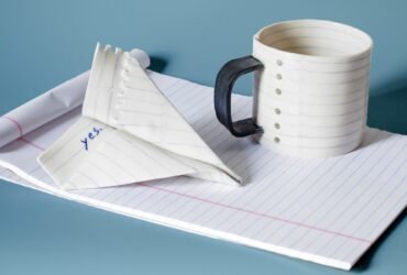 a mug and paper airplane sculpture that appear to made of lined paper atop white notepad