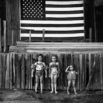 a black and white photo of three young girls in bathing suits standing in front of a wodoen stage with an american flag in background