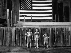 a black and white photo of three young girls in bathing suits standing in front of a wodoen stage with an american flag in background