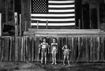 a black and white photo of three young girls in bathing suits standing in front of a wodoen stage with an american flag in background