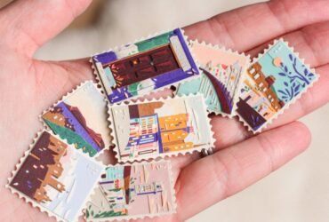 a hand holds up a collection of small, vibrant collages, each shaped like a stamp. they depict scenes with a building, water, architecture, and patterned designs