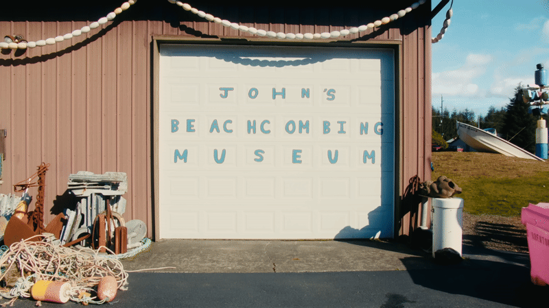 a gif from a short documentary showing a man wearing a black hat and jeans as he walks to the front of a garage door painted with the words "John's Beachcombing Museum"