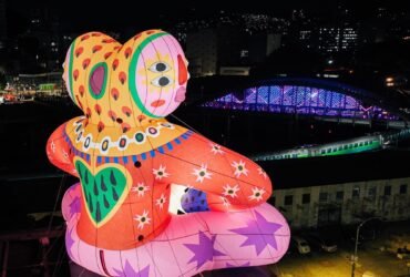 an aerial view of the rear of an inflatable public installation of a fantastical, brightly-colored figure perched on the edge of a city building, illuminated at night