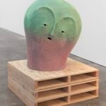 a large ceramic sculpture of a an abstract face in a green-to-pink gradient, installed on a pallet-inspired wood pedestal