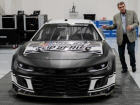 a car with "NASCAR" on hood parked inside a facility with man standing outside of car