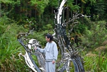 an Asian man stands at the base of a large, chrome-colored sculpture resembling a liquid splash frozen in time, installed in a field, with woods in the background