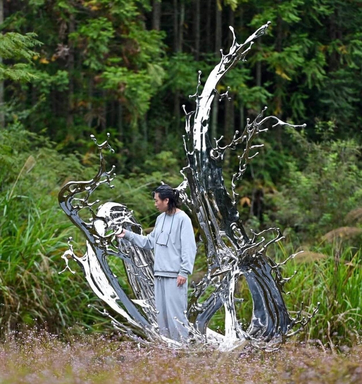 an Asian man stands at the base of a large, chrome-colored sculpture resembling a liquid splash frozen in time, installed in a field, with woods in the background