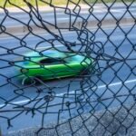 a photograph of a green car speeding along a road, seen through a chainlink fence that appears to be moving with the car