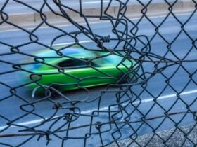 a photograph of a green car speeding along a road, seen through a chainlink fence that appears to be moving with the car