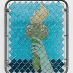 a wall sculpture of stained glass depicting the arm of the Statue of Liberty, installed behind a chainlink gate