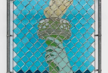 a wall sculpture of stained glass depicting the arm of the Statue of Liberty, installed behind a chainlink gate