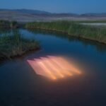 a river landscape in a desert with a peach-colored geometric light projection in the surface of the water